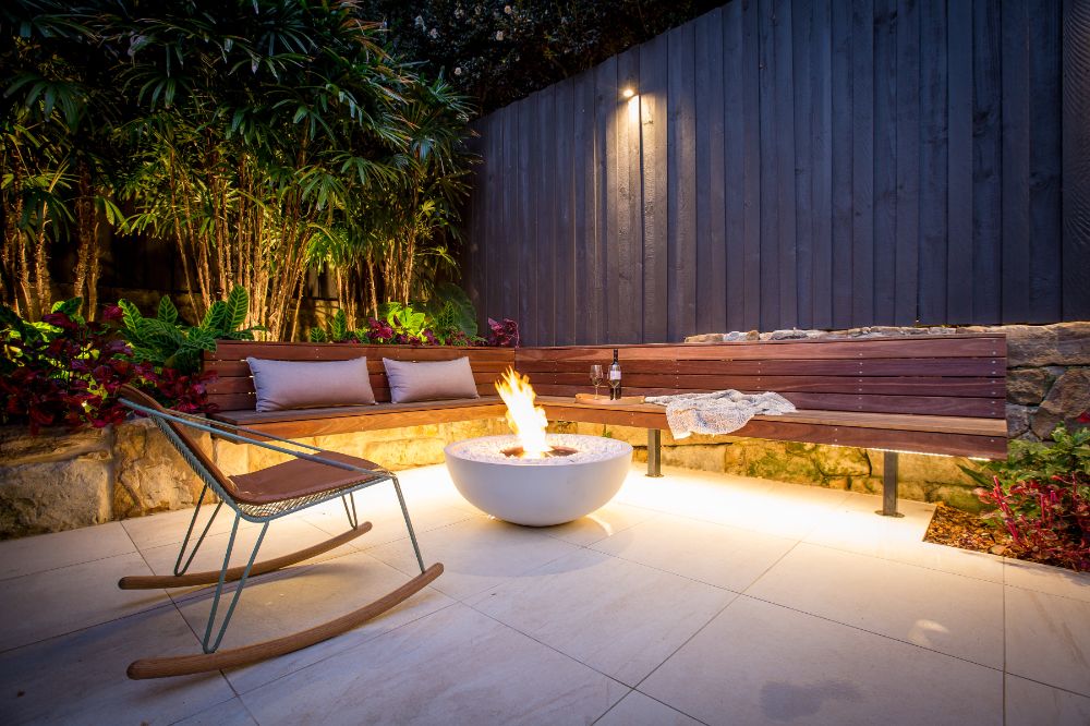 Outdoor patio with wooden bench, rocking chair, and fire pit at night.